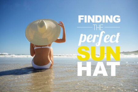 Back of a woman in a white bikini with a large sun hat sitting on the beach