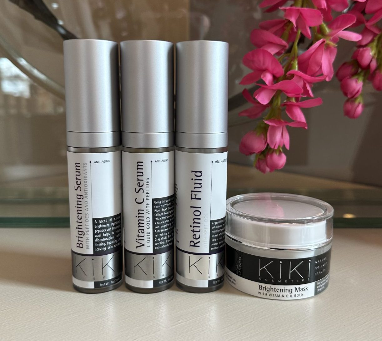 KIKI skincare collection featuring brightening serum, vitamin C serum, retinol fluid, and brightening mask with anti-aging benefits displayed with pink flowers.