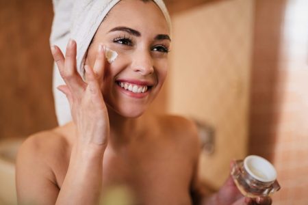 Woman with towel wrapped around head applying moisturizing cream to her face, holding cosmetic product jar.